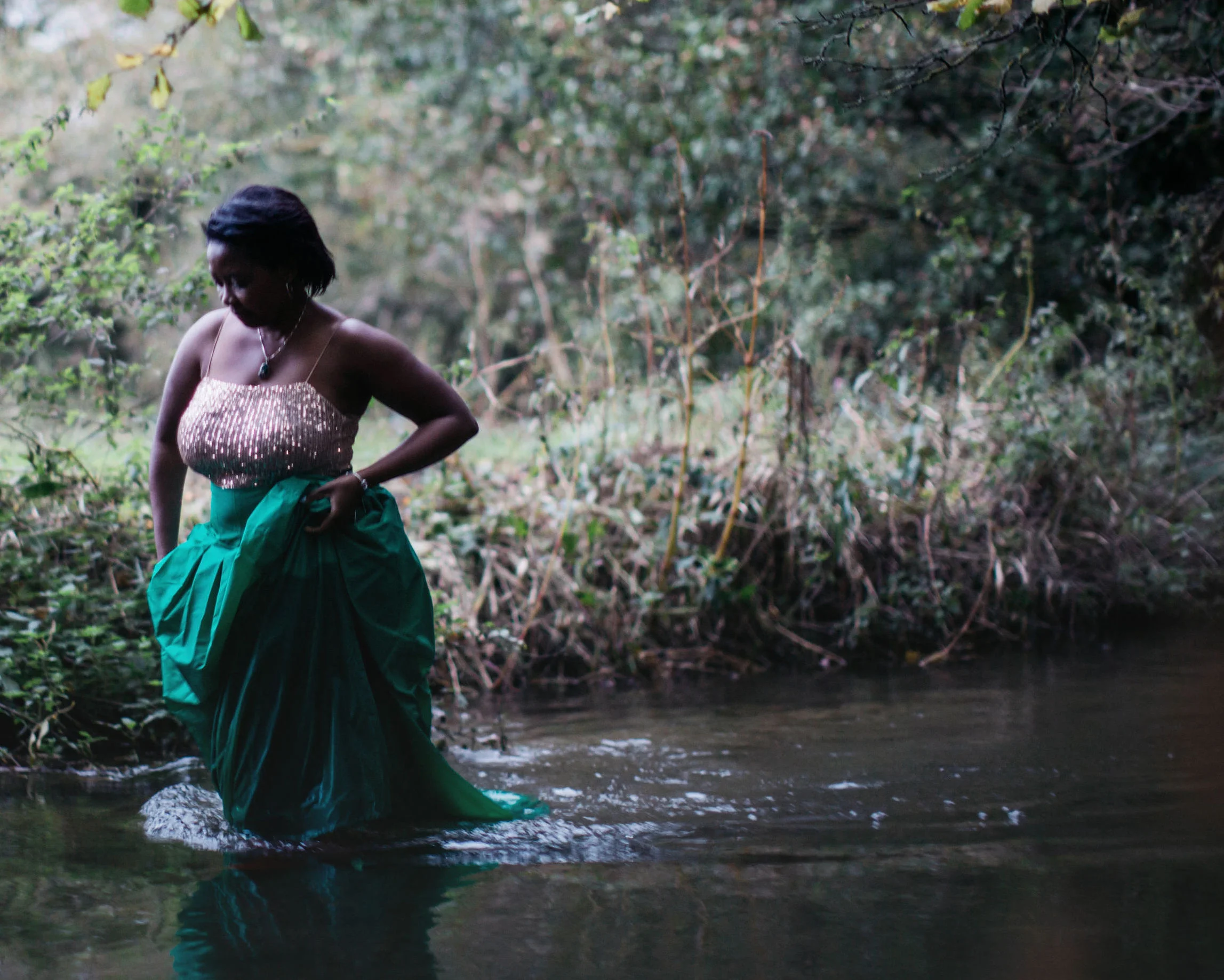 Folake standing in a woodland stream in a sequinned top and emerald skirt